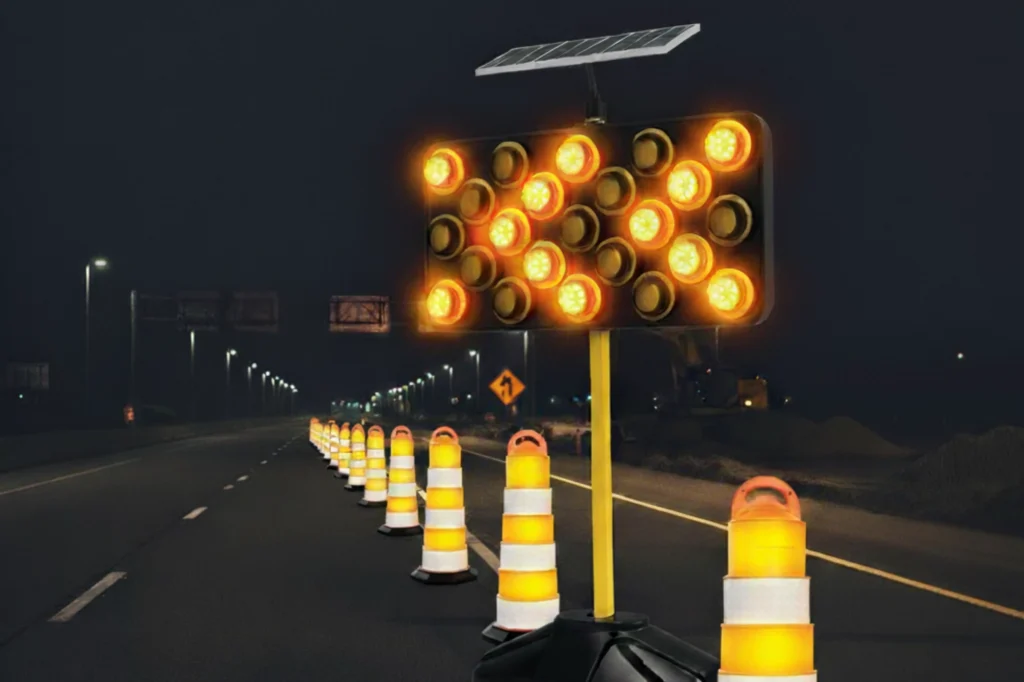 Flechero luminoso con panel solar y luces LED en modo clúster, instalado en una carretera nocturna junto a conos viales para señalizar un desvío y garantizar la seguridad vial.